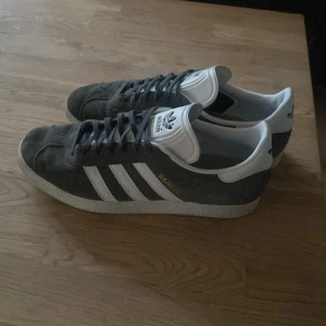 Adidas Gazelle i grå mocka - Säljer ett par klassiska Adidas Gazelle i grå mocka med vita detaljer och snörning. Skorna har den ikoniska tre-randiga designen på sidorna och en vit sula. Perfekta för en stilren outfit. Använda enbart ett fåtal gånger och priset är inte hugget i sten🤝