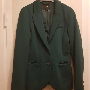 Mörk mint grön kavaj från H&M strl 36 - Snygg grön kavaj från H&M i storlek 36. Tillverkad av en blandning av polyester, viskos och elastan för en bekväm passform. Kavajen har långa ärmar och knappar framtill. Perfekt för att ge en stilren look.