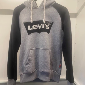 Grå och svart hoodie från Levi's - Säljer en snygg grå och svart hoodie från Levi's med en stir logga i fram. Tröjan har en stor ficka framtill och justerbar huva med snören. Perfekt för en avslappnad stil! 🖤