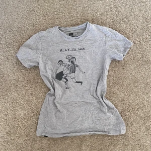 Grå t-shirt från Dedicated - Säljer en grå t-shirt från Dedicated med trycket 'Play to Win' och en tecknad fotbollsscen. Gjord av 100% ekologisk och Fairtrade-certifierad bomull. 