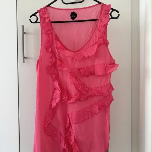 Rosa ärmlös topp med volanger - Säljer en söt rosa ärmlös topp från med volangdetaljer framtill. Perfekt för sommardagar och ger en lekfull touch till din outfit. Toppen har en luftig passform och är gjord i ett lätt material.