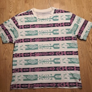 Mönstrad t-shirt från Levi's - Säljer en cool t-shirt från Levi's med ett unikt mönster i turkos, lila och vitt. T-shirten är kortärmad och har en normal passform. Perfekt för dig som vill ha en färgstark tröja till sommaren. Är väldigt bekväm och kostar ny 600 kr