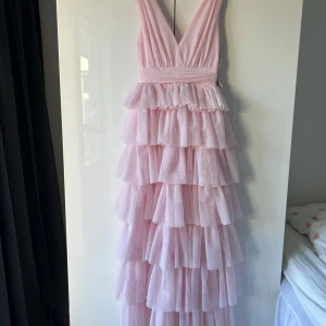 Ljusrosa volangklänning i tyll - Säljer en fantastisk ljusrosa långklänning i tyll med flera lager av volanger. Klänningen har en v-ringad design och är ärmlös. Har aldrig använts, perfekt till studentbal eller andra fina tillställningar🩷Jag är 161 och klänningen går ner till marken. 