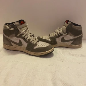 Nike Air Jordans i grått och vitt - Snygga Nike Air Jordans i en stilren kombination av grått och vitt. Skorna har en klassisk hög design med snörning och en rund tå. Perfekta för dig som vill ha en ikonisk sneaker med en tidlös look.