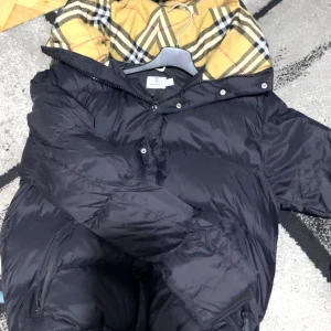 Svart dunjacka med mönstrad huva - Säljer en svart dunjacka med en snygg mönstrad insida i huvan. Jackan har en dragkedja och knappar framtill för extra skydd mot kyla. Perfekt för kalla dagar. Burberry jacka pris kan diskuteras 