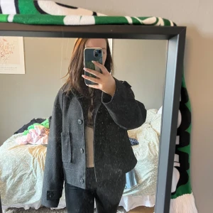 Mörkgrå kappa - Säljer en stilren mörkgrå kappa med knappar framtill. Jackan har en klassisk design med långa ärmar och en rak passform. Perfekt för kyligare dagar.