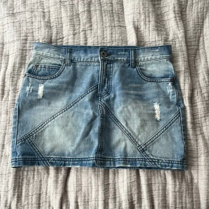 Blå jeanskjol med slitningar - Snygg blå jeanskjol med slitna detaljer och dekorativa sömmar. Kjolen har en klassisk femficksdesign och stängs med knapp och dragkedja. Säljer eftersom den är för liten för mig.