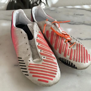 Adidas Predator fotbollsskor - Säljer ett par Adidas Predator fotbollsskor i vitt och rosa med svarta detaljer. Skorna har snörning och en rund tå. Perfekta för fotbollsspelare som vill ha stil och funktionalitet på planen.
