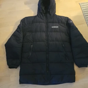 Svart dunjacka från Napapijri - Säljer en svart dunjacka från Napapijri med huva och dragkedja. Jackan har ett märke på ärmen och två fickor med dragkedja framtill. Perfekt för kalla dagar.Nästan ny.