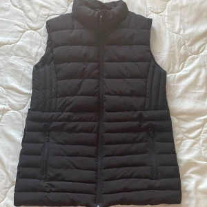 Svart dunväst från Mango - Snygg svart dunväst från Nine West med dragkedja framtill och två praktiska fickor med dragkedjor. Perfekt för lager-på-lager under kyliga dagar. Västen har en stilren design med en hög krage för extra värme.