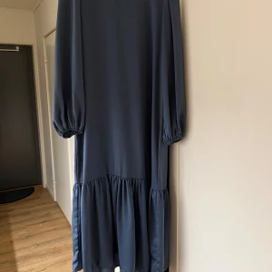 Mörkblå långklänning - Elegant havsblå långklänning med långa ärmar och volangdetalj vid fållen. Perfekt för en stilren look.