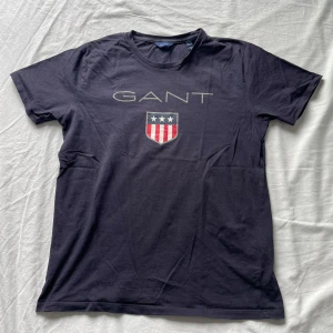 Svart/mörkblå t-shirt från GANT - Säljer en stilren svart t-shirt från GANT med ett tryck av deras logga och ett sköldmärke på framsidan. Perfekt för en casual look. T-shirten är kortärmad och gjord i mjukt bomullsmaterial.