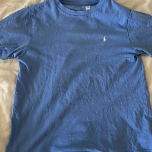 Blå t-shirt från Ralph Lauren - Säljer en snygg blå t-shirt från Ralph Lauren med klassisk logga på bröstet och stort tryck på ryggen. Perfekt för en avslappnad stil. Tillverkad i mjuk bomull för extra komfort. Svin fet till sommaren. Stolek M i barn.