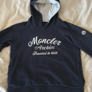 Mörkblå hoodie från Moncler - Säljer en snygg mörkblå hoodie från Moncler med texten 'Moncler Archive Founded in 1952' broderad på framsidan. Hoodien har en klassisk design med en justerbar huva och Monclers logotyp på ärmen. Perfekt för en stilren och bekväm look. Passar från 152-164, svin fet till våren och kalla sommardagar. Om ni vill ha kvitto hör av er. Fickor på båda sidor.
