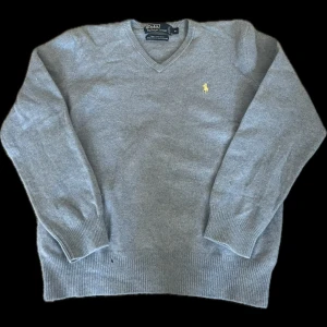 Grå ulltröja från Polo Ralph Lauren - Säljer en stilren grå tröja från Polo Ralph Lauren i 100% lammull. Tröjan har en v-ringad design med ett broderat gult logotypmärke på bröstet. Perfekt för kyligare dagar med sin mjuka och varma känsla. Storlek XL barnstorlek S i herr