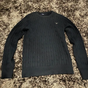 Svart kabelstickad tröja från Gant - Säljer en stilren marinblå kabelstickad tröja från Gant. Tröjan har en rund halsringning och långa ärmar, perfekt för kyligare dagar. Den har en diskret logga på bröstet. Passar både till jeans och chinos. Passar 159 cm