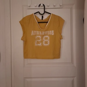 Gul croppad topp från H&M - Säljer en gul croppad topp från H&M med texten 'ATHLETICS 28' i vitt framtill. Toppen har korta ärmar och en vit kant vid halsringningen. Perfekt för en avslappnad stil.
