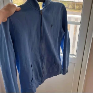 Blå hoodie från Ralph Lauren - Säljer en snygg blå hoodie från Ralph Lauren med dragkedja framtill. Tröjan har långa ärmar och en klassisk passform. Perfekt för en avslappnad stil.