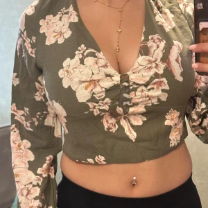 Grön blommig croppad blus med öppen rygg - Supersnygg grön croppad blus med stora ljusrosa blommor. Djup v-ringning framtill och öppen rygg med knytband upptill. Långärmad och figurnära passform, perfekt för dig som vill sticka ut lite extra. Tunt och skönt material.