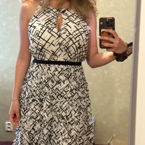 Vit och svart mönstrad klänning - Snygg vit kort klänning med svart grafiskt mönster och markerad midja med svart band. Klänningen har ärmlös halterneck-modell och en liten öppning framtill vid halsen. Tyget är lätt och luftigt, perfekt för varma dagar.