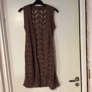 Brun lång stickad väst med knappar - Lång brun stickad väst från ginatricot med snyggt hålmönster och stora bruna knappar framtill. Västen är ärmlös och har en ribbad kant nedtill, perfekt att slänga över en t-shirt eller skjorta för en chill lager-på-lager-look.