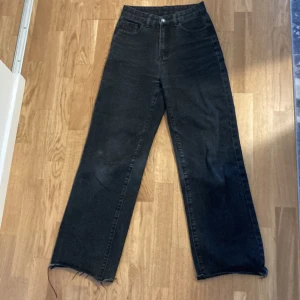 Svarta wide jeans med broderi - Säljer ett par svarta jeans med hög midja och raka, vida ben. Jeansen har råa, fransiga benslut och coolt vitt broderi i form av flames på bakfickorna. Perfekta för dig som vill ha en edgy vibe och gillar streetstyle.