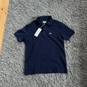 Marinblå pikétröja från Lacoste - Snygg marinblå pikétröja från Lacoste i 100% bomull. Klassisk design med krage, korta ärmar och den ikoniska gröna krokodilloggan på bröstet. Perfekt för dig som gillar stilrena och sportiga plagg. Passar dig som är ca 176 cm lång. Helt ny, precis köpt från Kidsbrandstore, ny pris 800, säljer för att det är för liten