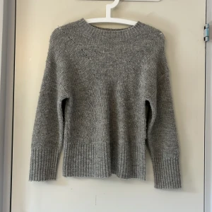 Grå stickad tröja från Lindex - Mysig grå stickad tröja från Lindex med bred ribbad mudd vid ärmslut och nederkant. Tröjan har en klassisk rund halsringning och lång ärm, perfekt för kyliga dagar. Enkel och stilren design som passar till det mesta.