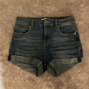 Mörkblå jeansshorts med uppvik - Snygga mörkblå jeansshorts med klassisk femficksdesign och uppvikta benslut. Shortsen har bälteshällor och en normal passform, perfekta för en chill sommarlook. Tillverkade i mjukt denimtyg som ger en avslappnad vibe.