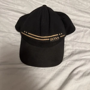 Svart keps från Hugo Boss - Snygg svart keps från Hugo Boss med guldfärgade detaljer och broderad logga framtill. Klassisk böjd skärm och stilren design som passar till det mesta. Tillverkad i bomull för en bekväm känsla. Skriv för mer bilder. 