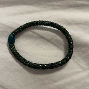 Glittrigt grönt armband med pärldetalj - Snyggt armband i mörkgrönt med glittrig yta och en blå pärldetalj. Perfekt accessoar för att ge din outfit lite extra bling. Elastiskt och lätt att ta på.