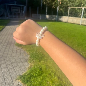 Pärlarmband med rosett och silverlås - Supergulligt armband med stora vita pärlor och en söt liten rosett som detalj. Armbandet har ett silvrigt lås som gör det enkelt att ta på och av. Perfekt accessoar för att lyfta din outfit med en klassisk och söt touch. Vi har en av denna sort.