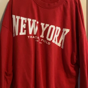 Röd sweatshirt med New York-tryck - Röd sweatshirt med stort vitt New York-tryck på bröstet och texten Track & Field under. Tröjan har rund halsringning och långa ärmar, perfekt för en sportig och avslappnad stil.