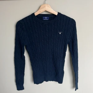 Marinblå kabelstickad tröja GANT XS - Kabelstickad marinblå tröja från GANT i stretchig bomull. Klassisk rund halsringning, diskret logga broderad på bröstet och ribbade muddar. Snygg passform som sitter tajt men bekvämt. Är något urtvättad 