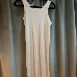 Vit ribbad långklänning - En enkel och stilren vit långklänning i ribbad bomull. Ärmlös modell med rund halsringning och figurnära passform. Perfekt för en avslappnad och trendig look.
