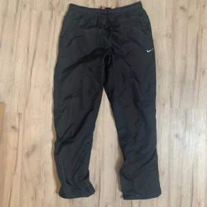 Svarta träningsbyxor från Nike - Svarta vintage träningsbyxor från Nike med klassisk Swoosh-logga på benet. Byxorna har elastisk midja med snörning och är tillverkade i lätt syntetmaterial som passar perfekt för träning. Sällsynta vintage byxor sitter löst perfekt för gym