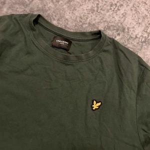Mörkgrön t-shirt från Lyle & Scott - Snygg mörkgrön t-shirt från Lyle & Scott med klassisk rund hals och korta ärmar. På bröstet sitter den ikoniska gula fågel-loggan. Tillverkad i mjuk bomull som är skön mot huden. Perfekt för en clean och stilren look.