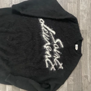 Saint Laurent svart mohairtröja - Svart långärmad stickad tröja från Saint Laurent med vit fuzzy logga framtill. Tröjan är gjord i mohairmix som ger en fluffig och lyxig känsla. Rund halsringning och relaxed fit, perfekt för sena sommarkvällar och sedan till hösten.