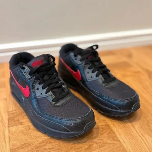 Nike Air Max svart/röd sneakers - Snygga Nike Air Max sneakers i svart med röda detaljer och klassisk röd swoosh. Skorna har svart mesh och skinn, samt gråa inslag vid snörningen. Luftbubblan i sulan ger extra komfort och cool look. Perfekta för dig som gillar streetstyle.