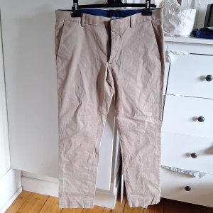 Beige chinos i bomull stretch W34 L30 - Snygga beige chinos med klassisk passform och raka ben. Byxorna har bälteshällor, dragkedja och knappstängning. Tillverkade i mjuk bomull med lite stretch för extra komfort. Perfekta till en avslappnad men stilren look.