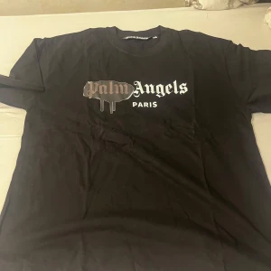 Svart Palm Angels t-shirt Paris L - Svart t-shirt från Palm Angels med ikoniskt tryck på bröstet där det står 'Palm Angels Paris' i vitt och brunt. Klassisk rund hals och korta ärmar. Skön passform och streetstyle-vibe, perfekt för dig som gillar trendiga märken.