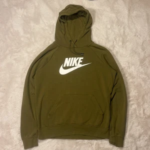 Olivgrön Nike hoodie  - Olivgrön hoodie från Nike. Tröjan har huva med snörning, känguruficka och långa ärmar. Perfekt för en avslappnad streetstyle-look. Tillverkad i mjuk bomullsmix som känns skön mot huden.
