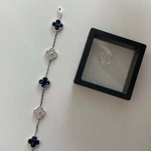 Silverfärgat armband med blommor - Stilrent armband i silverfärg med fem fyrklöverformade hängen, tre i mörkblå och två i glittrande silver. Kedjan är tunn och smidig, och armbandet har ett klassiskt lås. Perfekt accessoar för att lyfta din outfit.