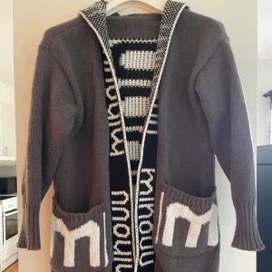 Grå stickad kofta med huva och mönster - Säljer en grå stickad kofta med huva och fickor framtill. Koftan har svart och vitt grafiskt mönster längs insidan och ryggen, samt textdetaljer. Perfekt för lager-på-lager och chill dagar.