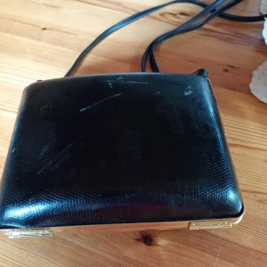 Svart axelväska i skinnimitation med gulddetaljer - Snygg svart axelväska i skinnimitation med guldfärgad ram och klassisk låsning. Väskan har en slät, lätt glansig yta med subtil struktur och en smal axelrem. Perfekt storlek för det viktigaste och ger en stilren vintagekänsla.