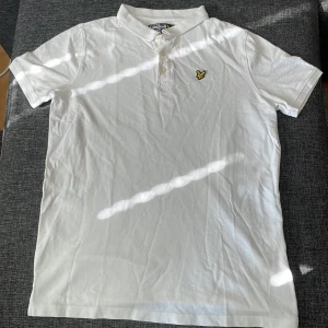 Vit pikétröja från Lyle & Scott - Klassisk vit pikétröja från Lyle & Scott med korta ärmar, krage och tre knappar framtill. Tröjan har det ikoniska gula fågel-loggan broderad på bröstet och är gjord i bomull för en skön känsla. Perfekt för en clean och stilren look. Och säljs pågrund av att den är för liten.