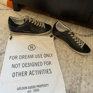 Svarta sneakers från Golden Goose - Snygga svarta sneakers från Golden Goose med coolt ormskinnsmönster och stjärndetalj på sidan. Skorna har vita snören, vit sula och svart häl. Materialet är skinn med textur och de har klassisk rund tå och platt sula. Perfekta för dig som gillar streetstyle.