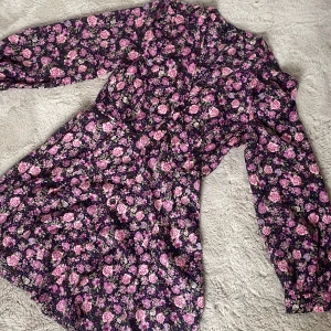 Blommig kortklänning med knappar - Supersöt kortklänning med lila och rosa blommor på mörk botten. Klänningen har långa ärmar, markerad midja (insydd) och knappar framtill. 