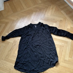 Svart långärmad klänning H&M - Svart långärmad klänning från H&M i glansigt, mjukt material. Klänningen har v-ringning, långa ärmar med breda manschetter och en lös, flowy passform. Perfekt för dig som gillar stilrena och enkla plagg med en touch av elegans.