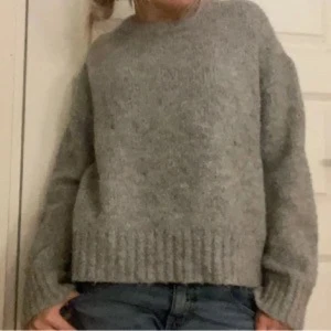 Grå stickad tröja med lång ärm - Mysig grå stickad tröja med rund hals och långa ärmar. Tröjan har en ribbad kant nedtill och vid ärmsluten, vilket ger en snygg och enkel look. Perfekt att slänga på sig när det är kyligt ute.